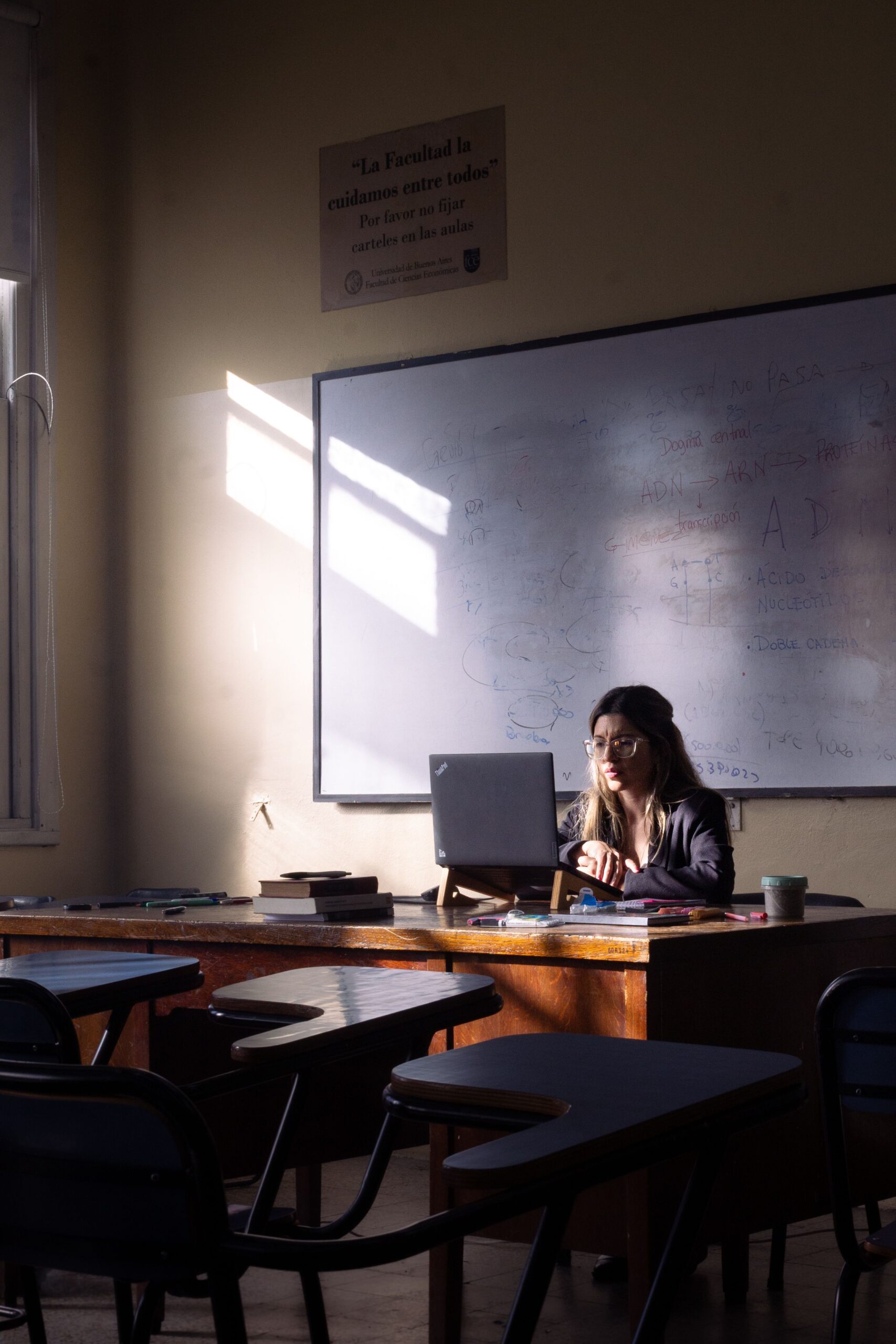 A female teacher focused on her work at a computer in a sunlit classroom with a whiteboard.